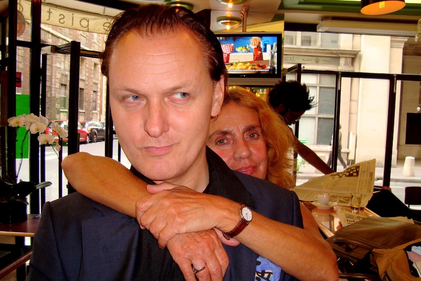 A man in a dark suit being hugged by a woman in a European cafe.