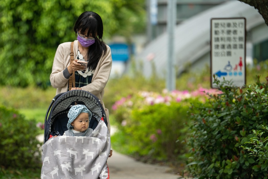 A woman wearing a purple mask looks at her phone as she leans on a stroller with her baby inside.