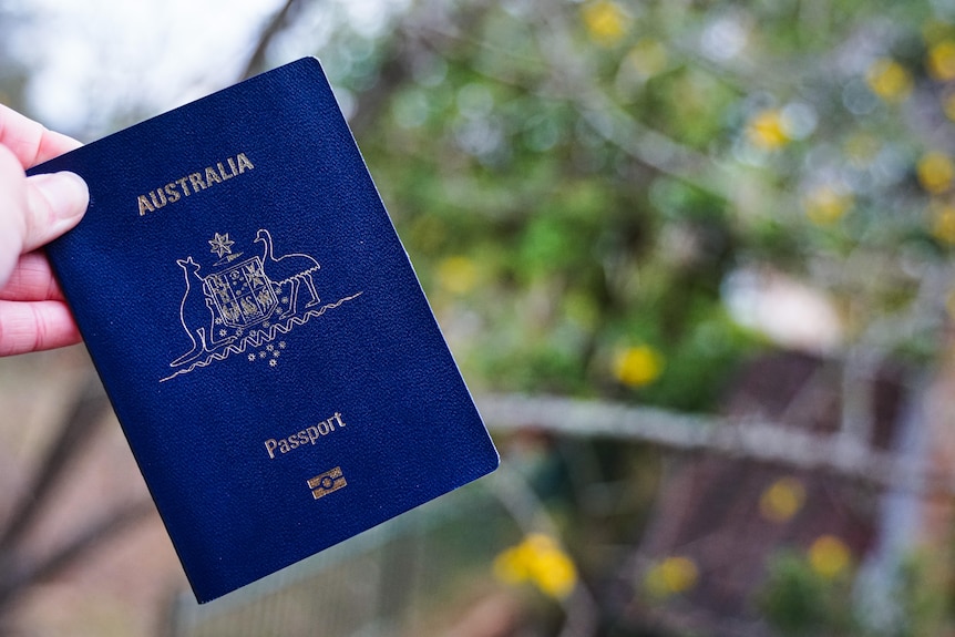Hand holding an Australian passport by its blue cover, with a garden in the background.