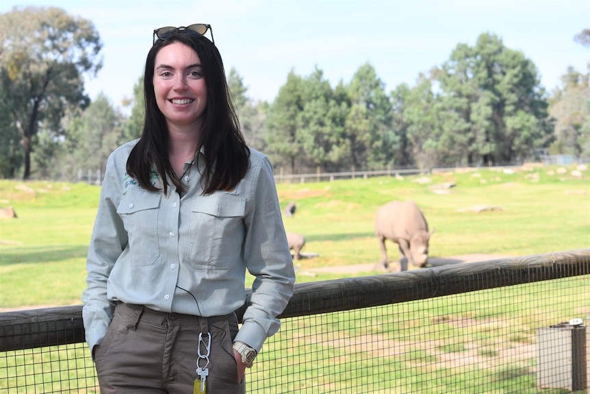 A lady with black hair smiles at the camera with two black rhinos behind her.