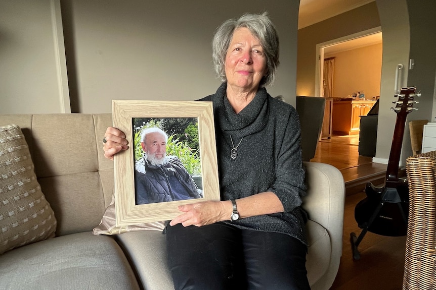 A woman with grey hair sitting on a sofa holding a framed photo of a man with white hair and a beard
