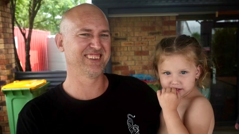 A smiling man in a dark shirt holding a little girl while he stands outside a house.