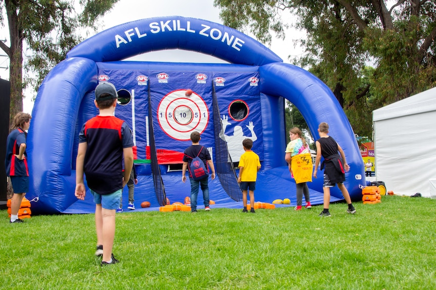 half a dozen children stand in front of an inflatable 'AFL skill zone', one has just kicked a football towards a target