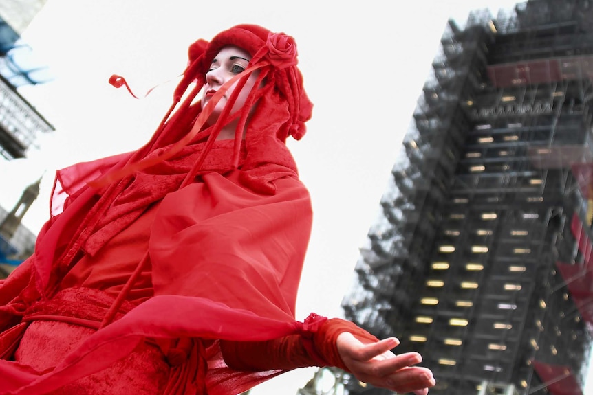 A woman has her face painted in white and is dressed head to toe in red flowing garments. She holds out her hands by her side.