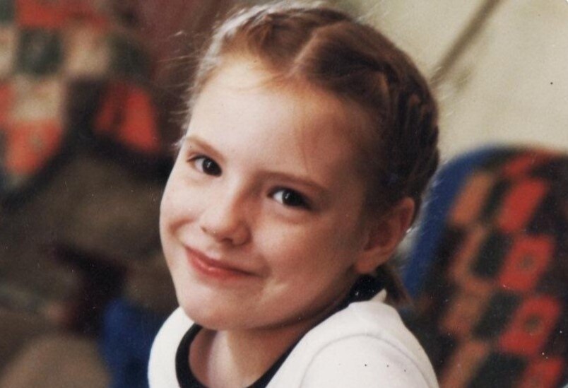 A young girl with blond hair and braids smiles at the camera