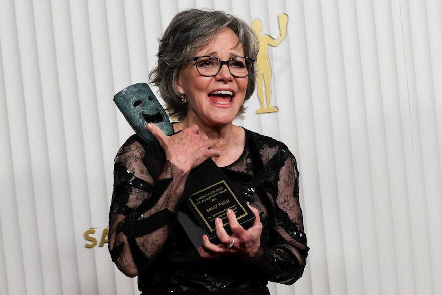 Sally Field sosteniendo un trofeo en su pecho con la boca abierta y anteojos para leer