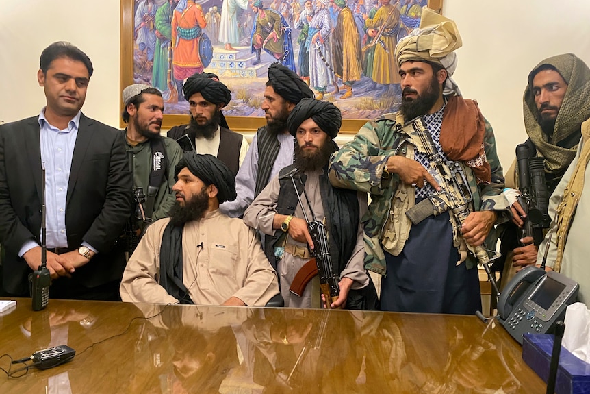 Armed men in traditional Afghan clothing stand around a man seated at a timber desk.