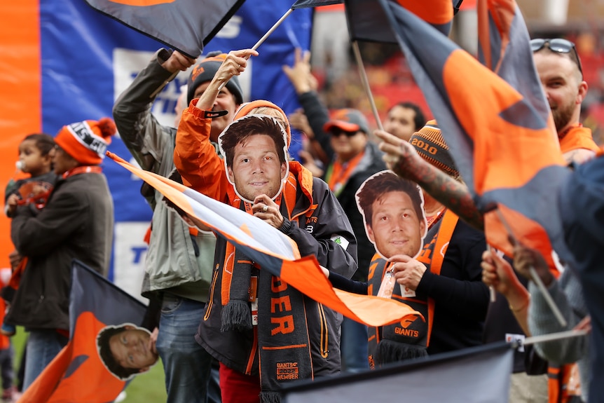 A crowd of GWS AFL fans stand in the crowd, with two of them holding Toby Greene masks in front of their faces. 