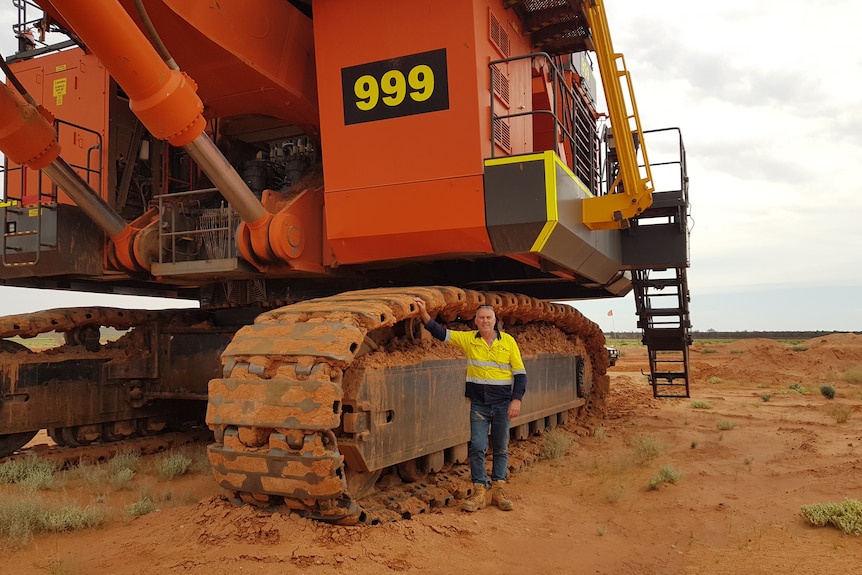 A man in high-vis stands in front of an enormous excavator.