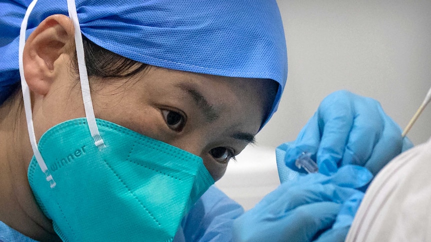 A woman in a blue cap and face mask holds a needle to someone's shoulder