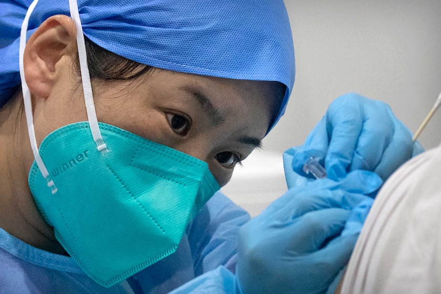 Une femme avec une casquette bleue et un masque facial tient une aiguille sur l'épaule de quelqu'un