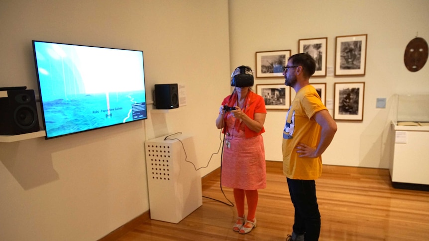 A woman stands with a man while wearing the virtual reality headset in front of a screen.