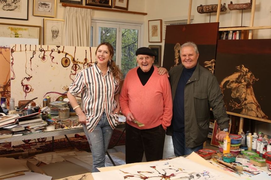 John Olsen smiling, standing between his daughter and son, art all around them.