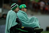 Two cricket players on the sidelines wrapped up in blankets