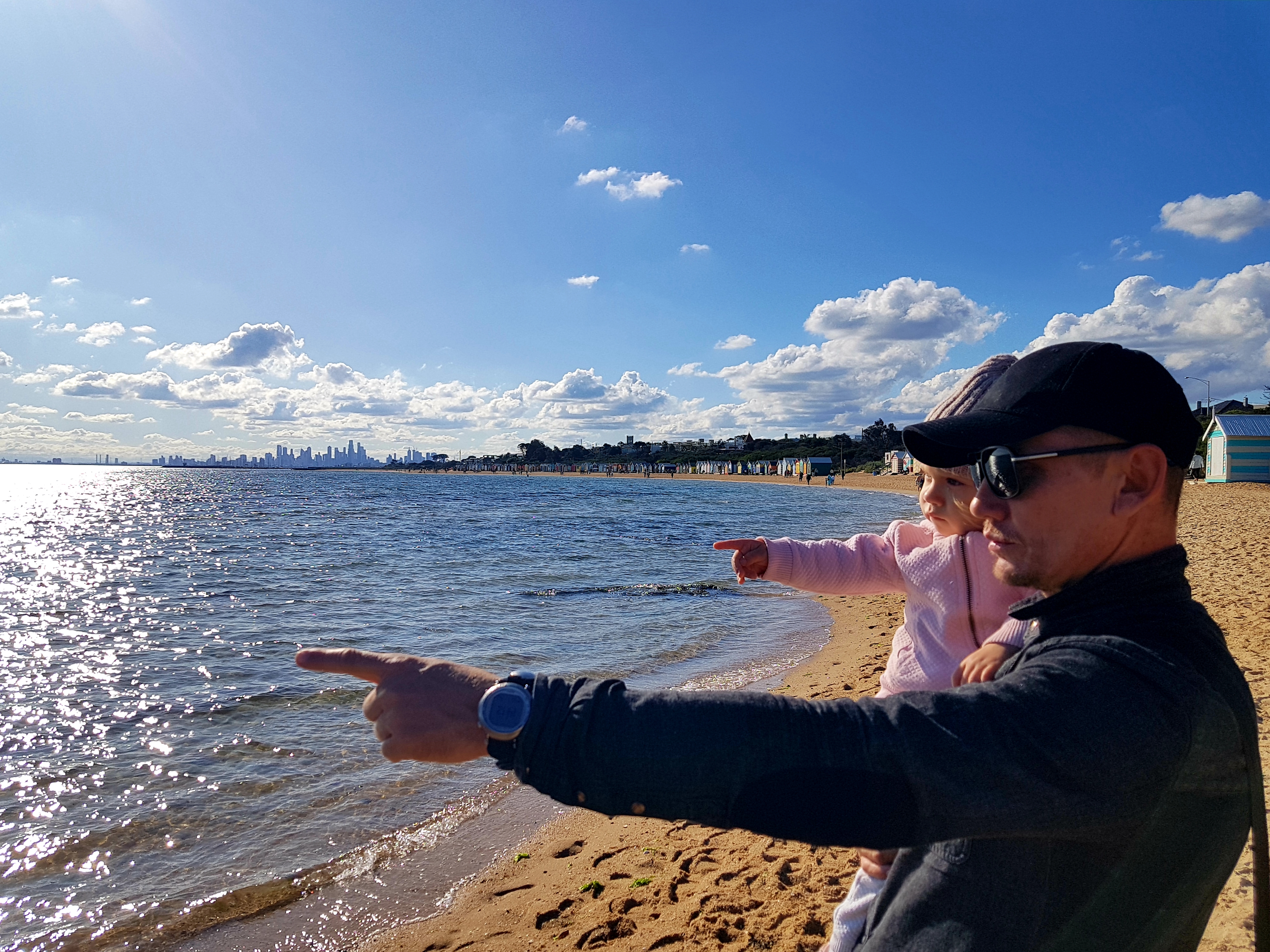 A man holds his young daughter, they are on the beach pointing to the waves