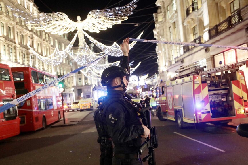 An armed police officer ducks under police tape on Oxford Street, London. Christmas lights overhead.