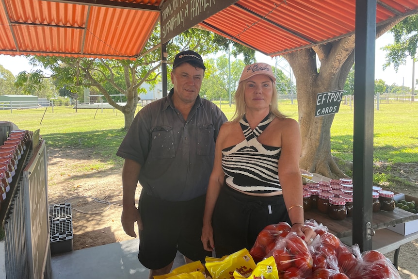 Man with old workshirt on stands next to a woman with a pink hat and black and white halter neck top. 