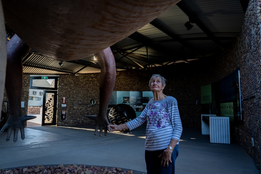 An elderly woman stands underneath the belly of a lifesize dinosaur statue and holds its hand.