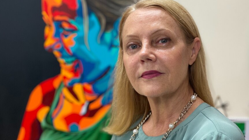 A woman with shoulder-length blonde hair stands in front of a colourful painting.