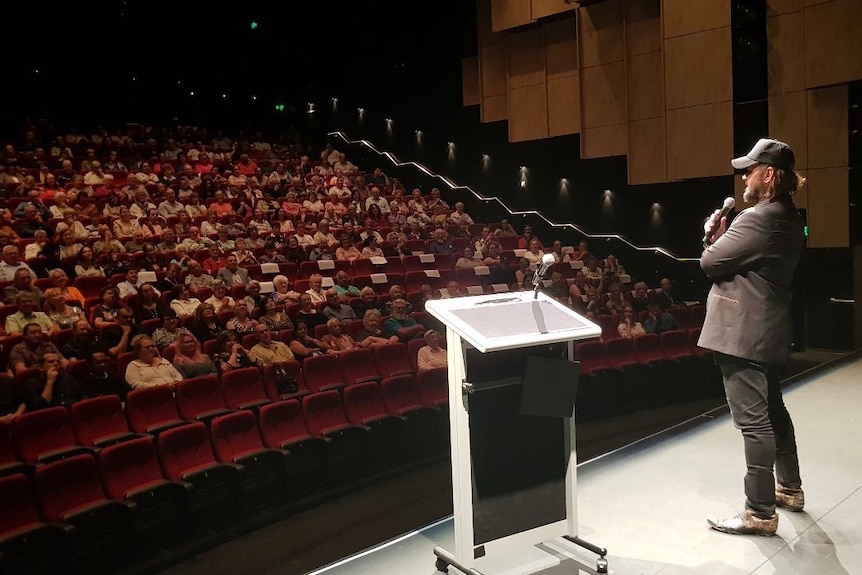 Film director Warwick Thornton stands on stage with microphone and podium addressing audience inside cinema.