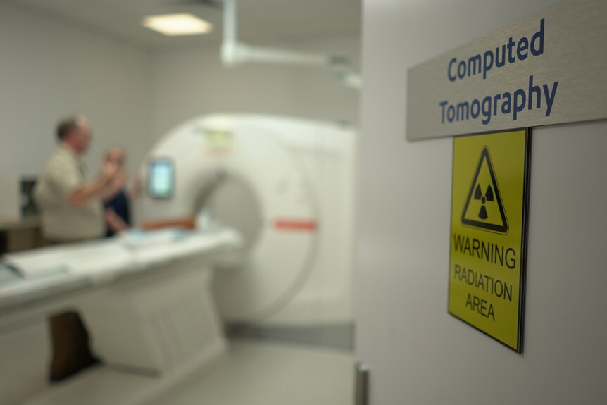A close-up of a computed tomography sign, with a man and woman in the background looking at the CT scanner