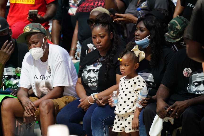 Four African American people sit solemnly at front of colourful crowd, with some wearing face masks.