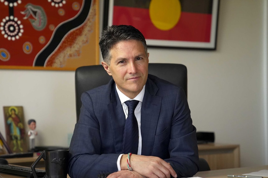 Man in suit and tie with arms resting on wooden desk in office with artwork behind him