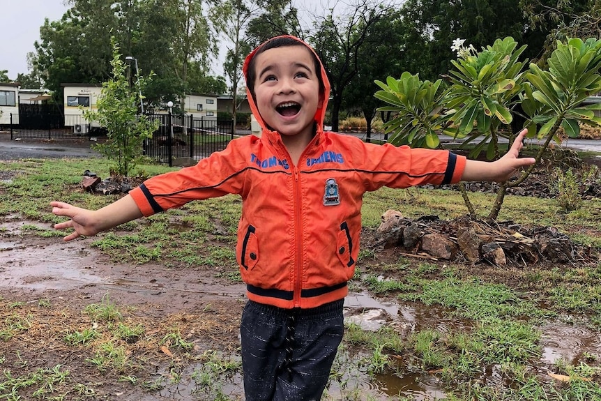 A young boy wearing an orange sweater smiles with arms wide in the bush