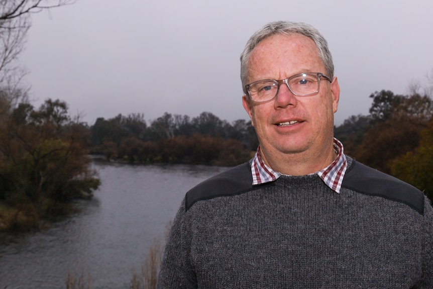 A man wearing a grey jumper and collared shirt, as well as glasses, stands in front of a river.