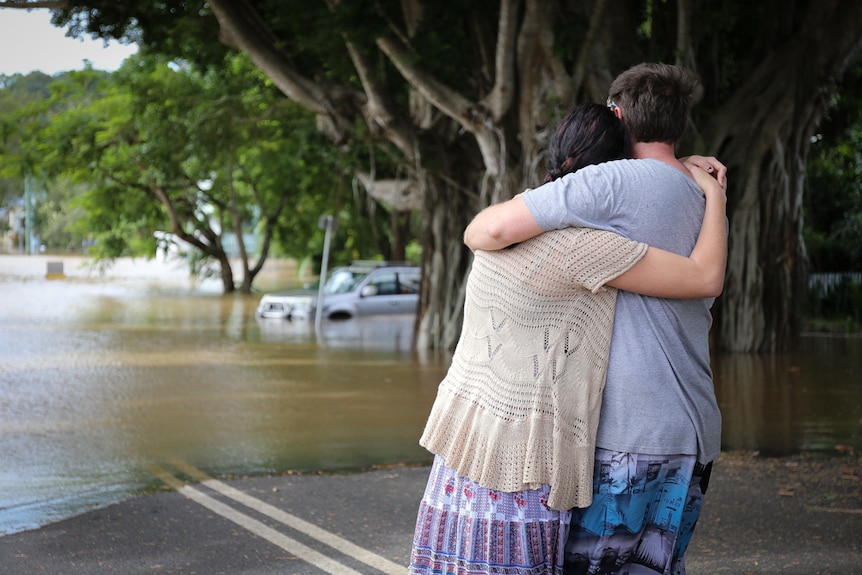 Couple hugging with car submerged in flood waters in the distance