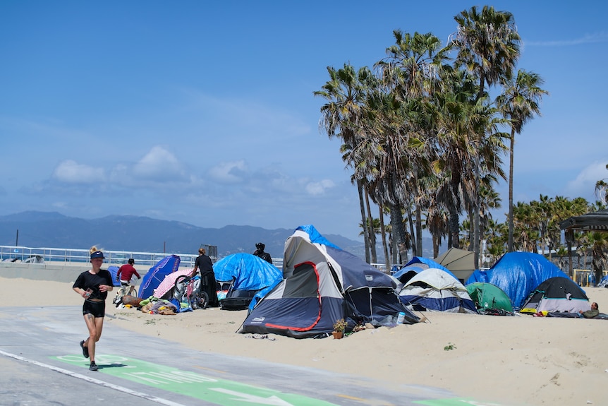 A woman jogs past a row of tents lined along a beach surrounded by palm trees 