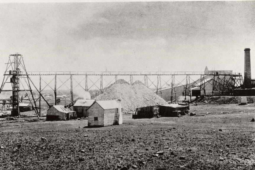 A black-and-white photo of huts, a large mound, and a long horizontal industrial structure.