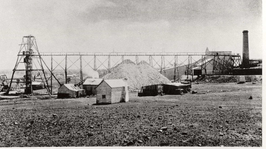 A black-and-white photo of huts, a large mound, and a long horizontal industrial structure.