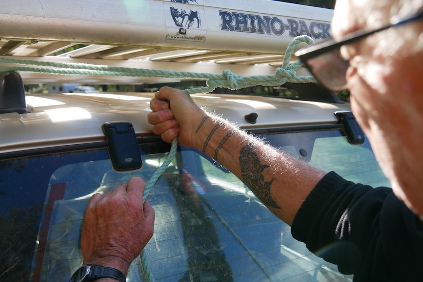 Close-up of a man pulling a rope tight on roof-racks, tattoos on his arm.