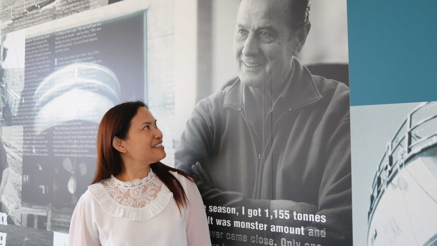 A dark-haired woman smiles at a large wall-mounted profile picture of a middle-aged man