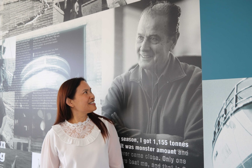 A dark-haired woman smiles at a large wall-mounted profile picture of a middle-aged man