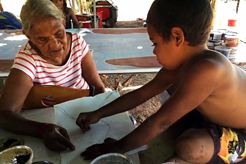 A child sits on a table and draws as an elderly woman looks on.