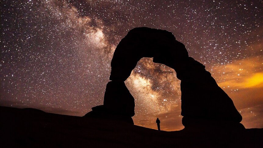 Person staring at starry night sky under a curved rock formation