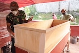 Two men standing on either side of a plywood coffin, with other coffins wrapped in plastic behind them.