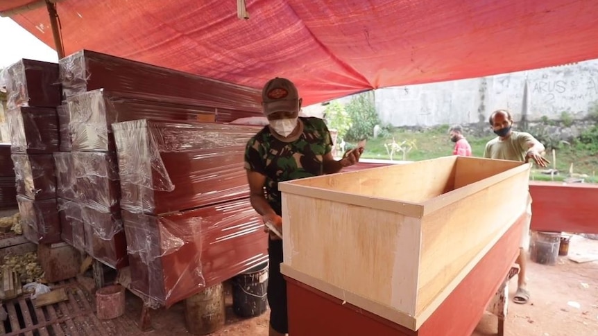 Two men standing on either side of a plywood coffin, with other coffins wrapped in plastic behind them.