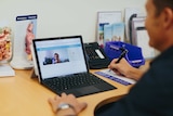 Man sits at desk looking at laptop screen, woman is displayed on screen