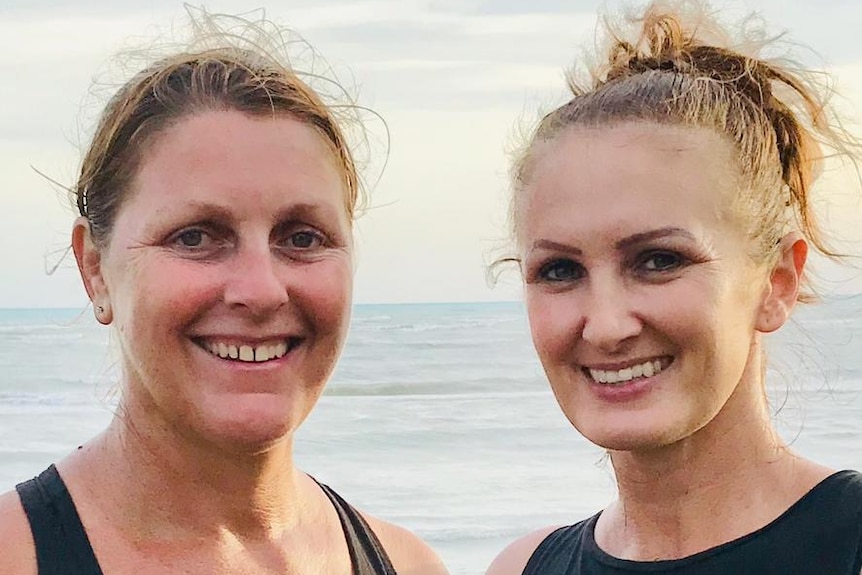 Two women stand on a beach smiling.