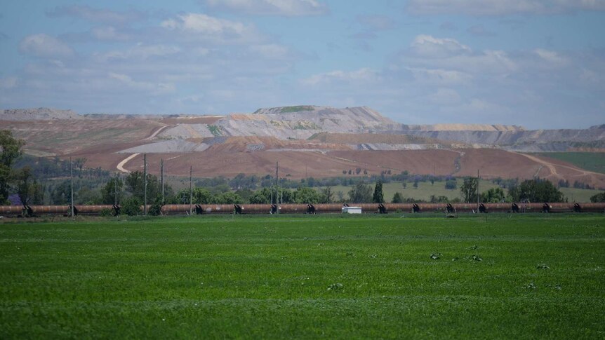 A train goes through a green paddock in front of an open cut coal mine