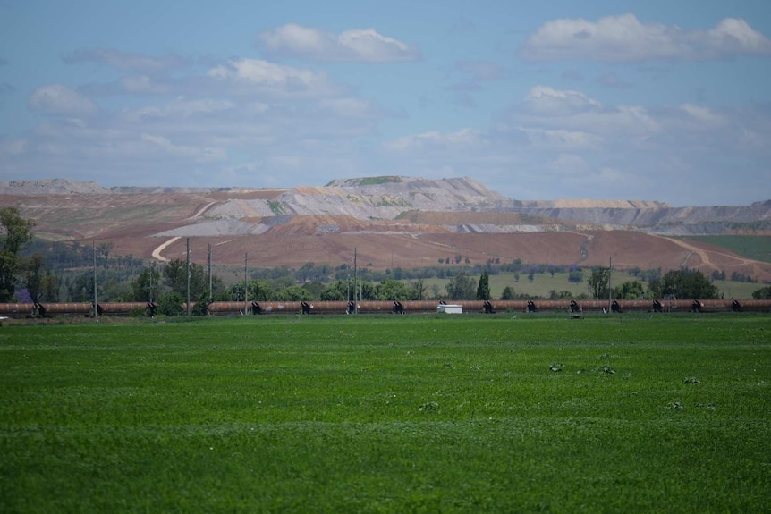 A train goes through a green paddock in front of an open cut coal mine