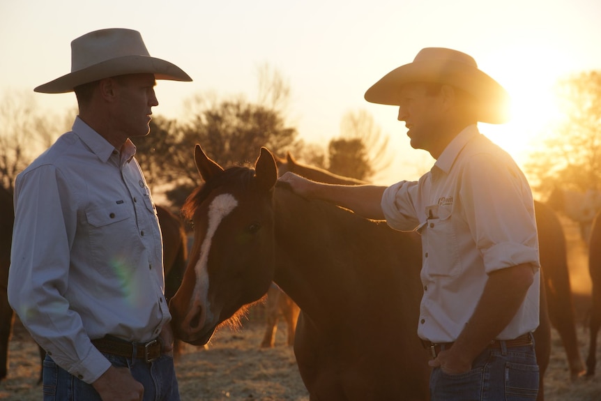Two men wearing cowboy hats stand next to a horse at sunrise.