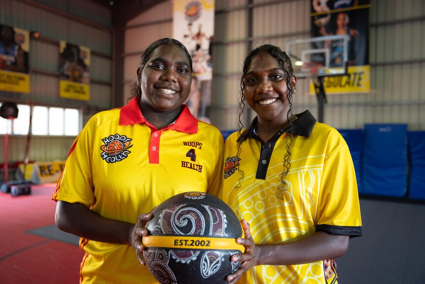 Two Indigenous teenage girls in yellow basketball jerseys holding a basketball, smiling at the camera, on an indoor court.