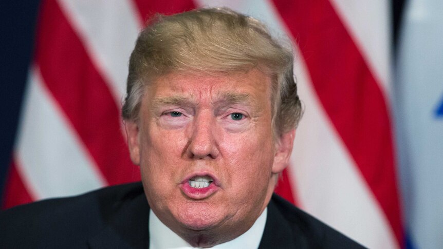 Donald Trump sits in front of an American flag.