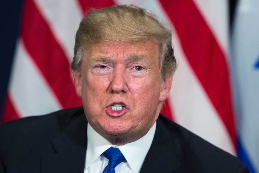 Donald Trump sits in front of an American flag.