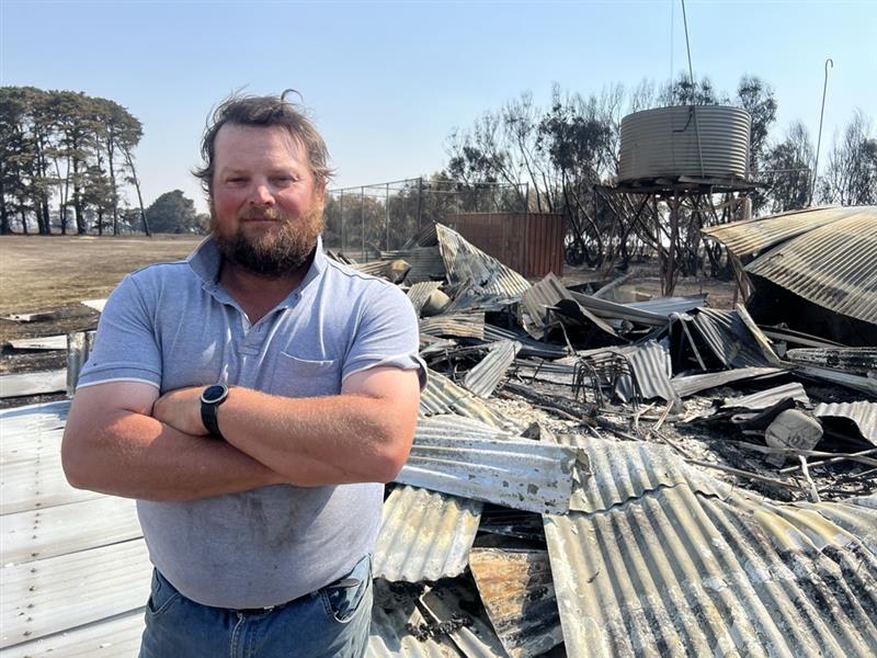 Un hombre con polo, vaqueros y barba se encuentra con los brazos cruzados frente a los escombros quemados. 