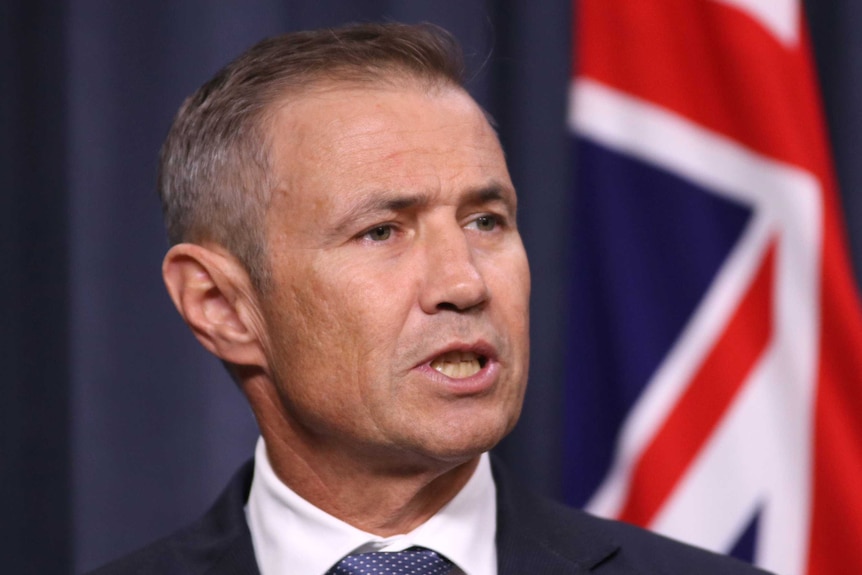 A tight head and shoulders shot of WA Health Minister Roger Cook standing in front of a WA flag.